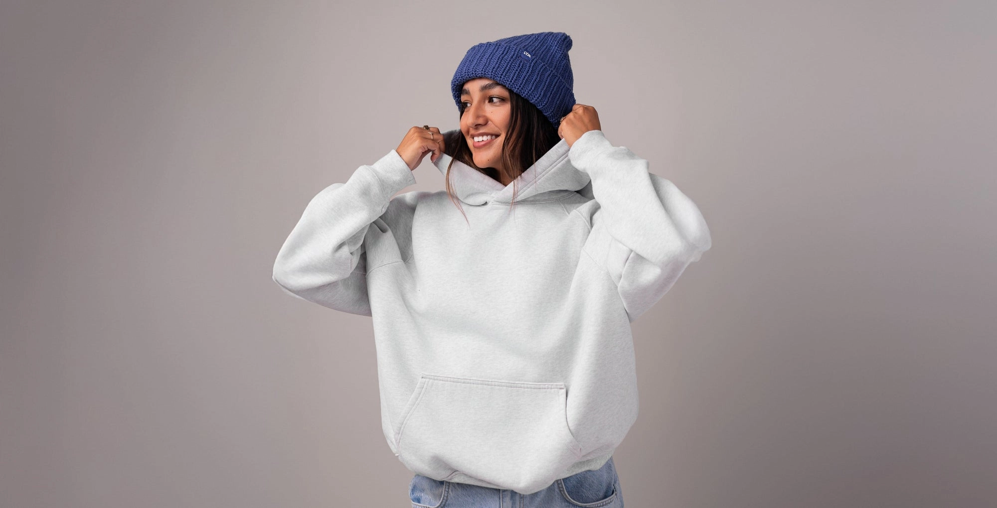 A woman in a blue Essentials beanie pulling up the hood of a gray sweatshirt