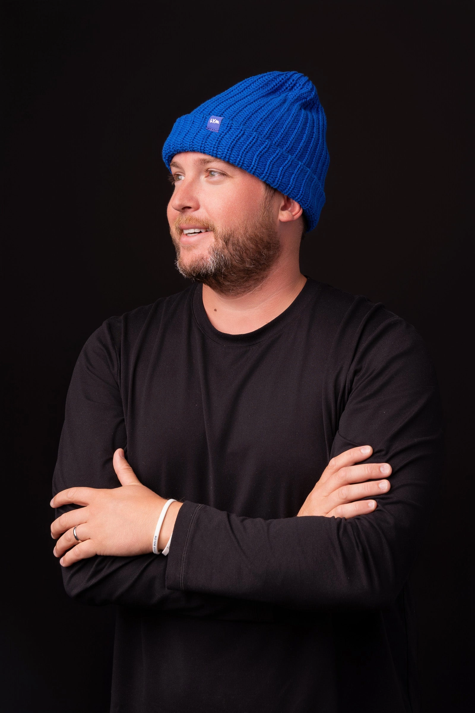 A man with crossed arms in a bright blue LYM beanie on a black background