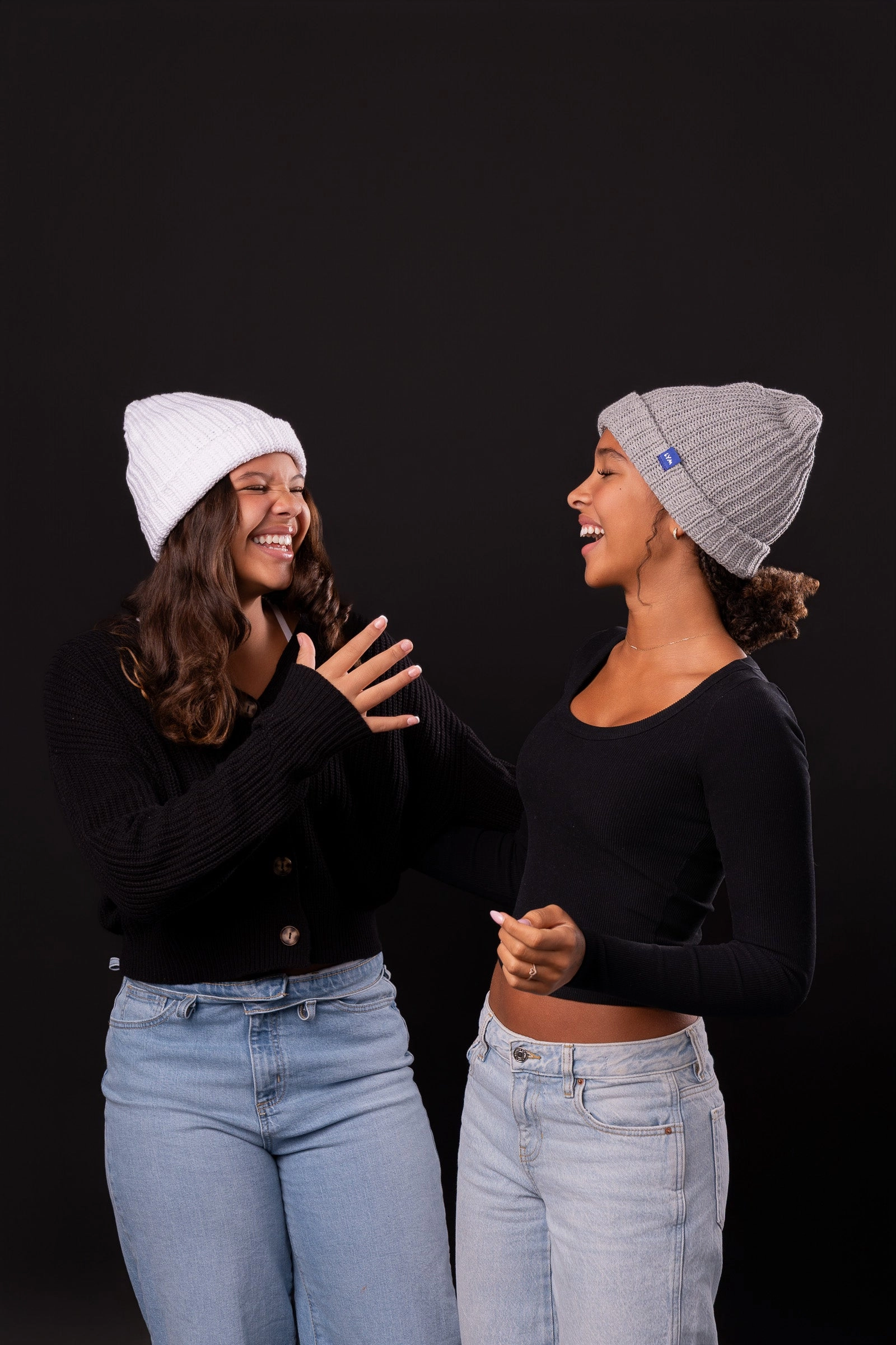Two friends laughing in white and gray LYM beanies on a black background