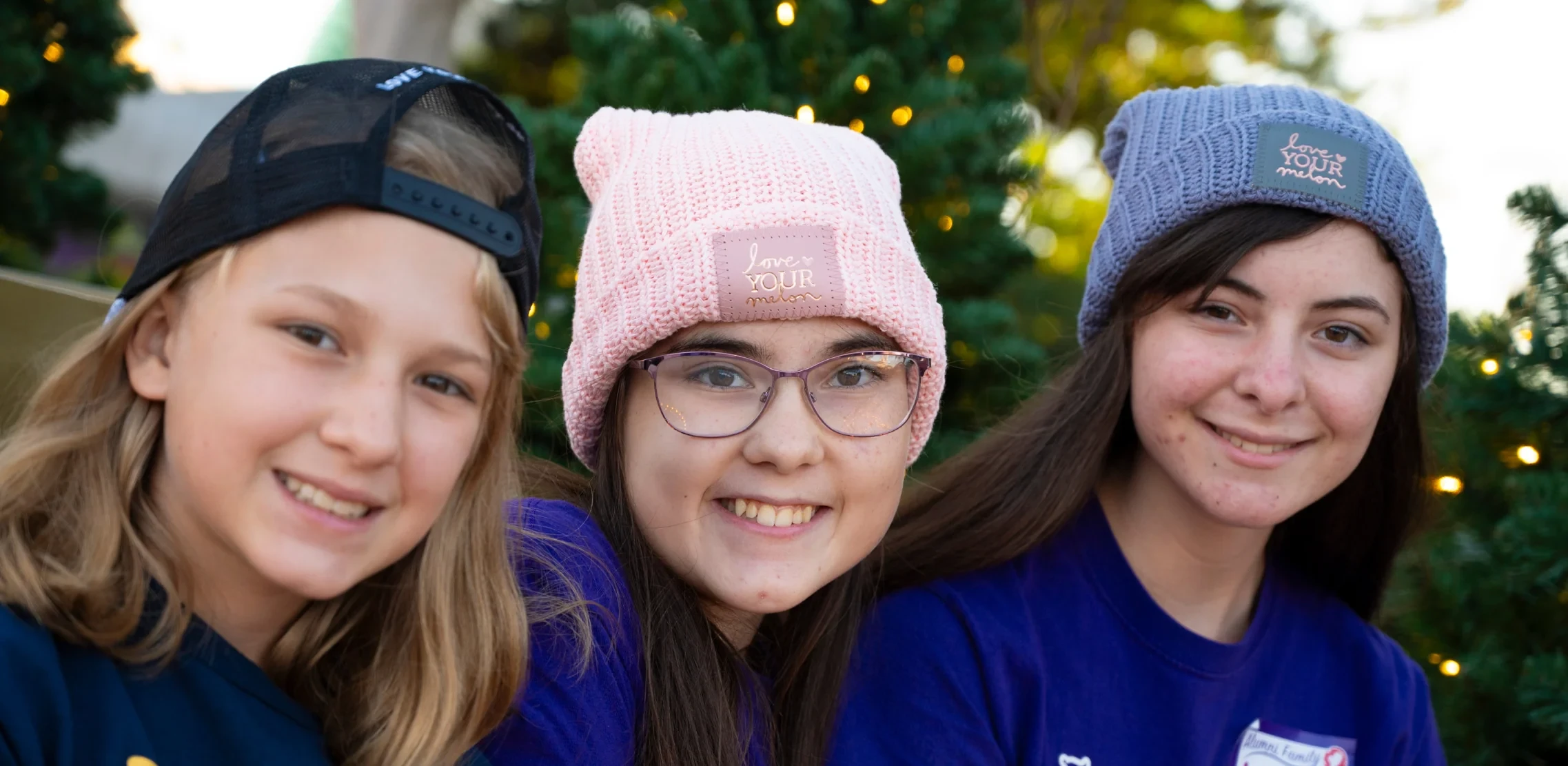 Three girls smiling in a black cap, a pink beanie and a lavender beanie with the Love Your Melon patch